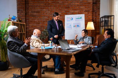 Hintli iş adamları projeyi tartışırken sunucu ayakta duruyor. Hintli iş adamı kıdemli yönetici ekibine stratejiyi açıklıyor.