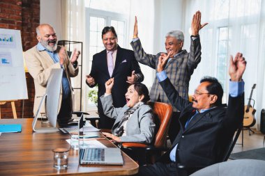 Hintli beş iş adamı laptopta çalışırken başarıyı kutluyor. Hintli üst düzey Asyalı işadamları modern ofiste masada otururken veya ayakta dururken kâr ve başarıyı alkışlıyor.