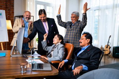 Hintli beş iş adamı laptopta çalışırken başarıyı kutluyor. Hintli üst düzey Asyalı işadamları modern ofiste masada otururken veya ayakta dururken kâr ve başarıyı alkışlıyor.