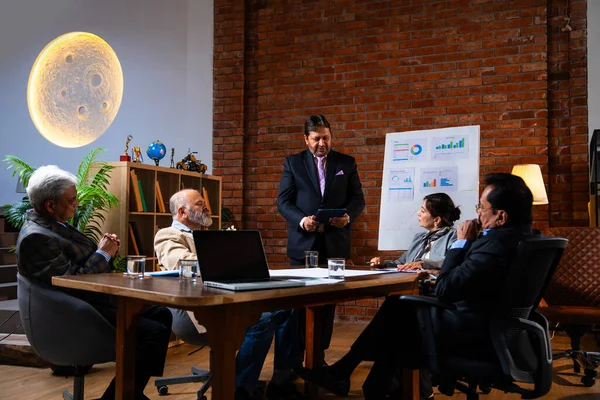 Hintli iş adamları projeyi tartışırken sunucu ayakta duruyor. Hintli iş adamı kıdemli yönetici ekibine stratejiyi açıklıyor.