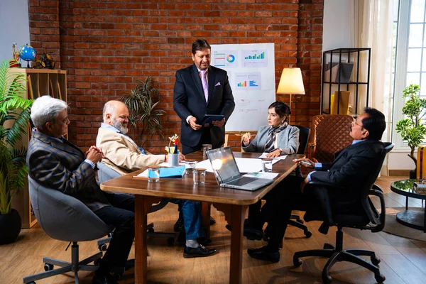 Hintli iş adamları projeyi tartışırken sunucu ayakta duruyor. Hintli iş adamı kıdemli yönetici ekibine stratejiyi açıklıyor.