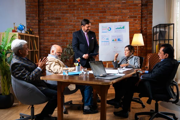 Hintli iş adamları projeyi tartışırken sunucu ayakta duruyor. Hintli iş adamı kıdemli yönetici ekibine stratejiyi açıklıyor.