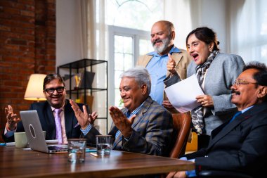 Hintli beş iş adamı laptopta çalışırken başarıyı kutluyor. Hintli üst düzey Asyalı işadamları modern ofiste masada otururken veya ayakta dururken kâr ve başarıyı alkışlıyor.