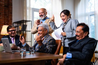 Hintli beş iş adamı laptopta çalışırken başarıyı kutluyor. Hintli üst düzey Asyalı işadamları modern ofiste masada otururken veya ayakta dururken kâr ve başarıyı alkışlıyor.