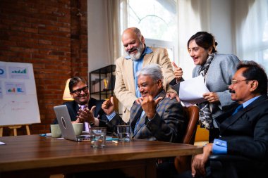 Hintli beş iş adamı laptopta çalışırken başarıyı kutluyor. Hintli üst düzey Asyalı işadamları modern ofiste masada otururken veya ayakta dururken kâr ve başarıyı alkışlıyor.