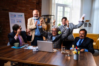 Hintli beş iş adamı laptopta çalışırken başarıyı kutluyor. Hintli üst düzey Asyalı işadamları modern ofiste masada otururken veya ayakta dururken kâr ve başarıyı alkışlıyor.