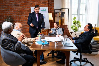 Hintli iş adamları projeyi tartışırken sunucu ayakta duruyor. Hintli iş adamı kıdemli yönetici ekibine stratejiyi açıklıyor.