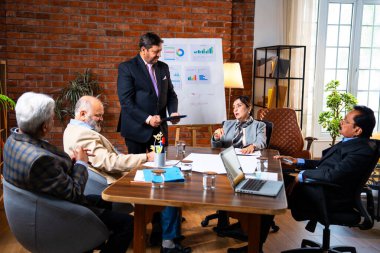 Hintli iş adamları projeyi tartışırken sunucu ayakta duruyor. Hintli iş adamı kıdemli yönetici ekibine stratejiyi açıklıyor.