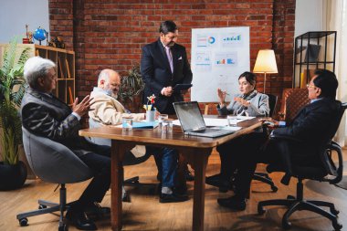 Hintli iş adamları projeyi tartışırken sunucu ayakta duruyor. Hintli iş adamı kıdemli yönetici ekibine stratejiyi açıklıyor.