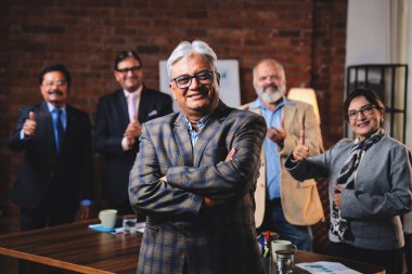 Hintli kıdemli iş adamının portresi Hintli üst düzey yönetici yönetici kameraya kendinden emin bir ifadeyle gülümserken meslektaşı modern kurumsal ofiste arkasında duruyor.