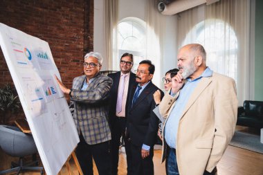 Hintli iş adamları grafikte projeksiyonu tartışırken, üst düzey Asyalı yöneticiler yakın durup modern kurumsal toplantı odasındaki beyaz panodaki büyüme verilerini gözden geçiriyorlar.