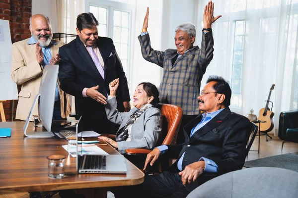 Hintli beş iş adamı laptopta çalışırken başarıyı kutluyor. Hintli üst düzey Asyalı işadamları modern ofiste masada otururken veya ayakta dururken kâr ve başarıyı alkışlıyor.
