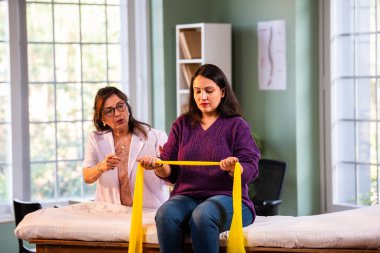 Fizyoterapist Hintli kadın hasta triseps ve omuz güçlendirme egzersizleri yapıyor modern klinikte direnç bandını kullanarak rehabilitasyon ve iyileşme için kıdemli rehberlik altında