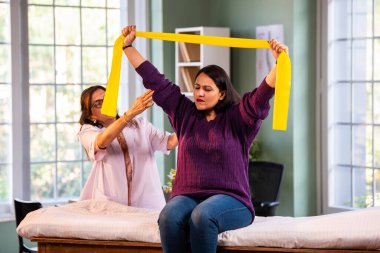 Fizyoterapist Hintli kadın hasta triseps ve omuz güçlendirme egzersizleri yapıyor modern klinikte direnç bandını kullanarak rehabilitasyon ve iyileşme için kıdemli rehberlik altında