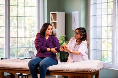 Hintli kadın fizyoterapist, modern klinikte yapılan detaylı istişare sırasında genç kadın hastaların bilek ağrılarını inceliyor, ortak değerlendirme ve rehabilitasyon alanındaki uzmanlığı vurguluyor.