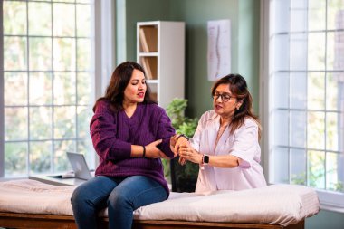 Hintli kadın fizyoterapist, modern klinikte yapılan detaylı istişare sırasında genç kadın hastaların bilek ağrılarını inceliyor, ortak değerlendirme ve rehabilitasyon alanındaki uzmanlığı vurguluyor.