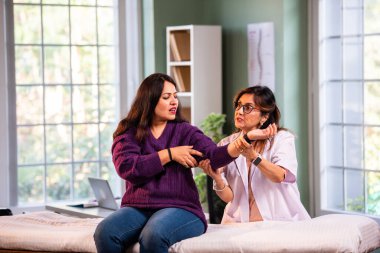 Hintli kadın fizyoterapist, modern klinikte detaylı klinik değerlendirmeler sırasında genç bir kadının dirsek ağrısını inceliyor, ortak bakım ve rehabilitasyon terapisinde uzman olduğunu vurguluyor.