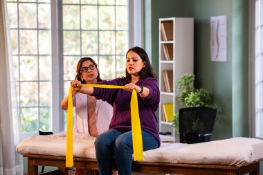 Fizyoterapist Hintli kadın hasta triseps ve omuz güçlendirme egzersizleri yapıyor modern klinikte direnç bandını kullanarak rehabilitasyon ve iyileşme için kıdemli rehberlik altında