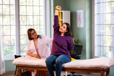 Fizyoterapist Hintli kadın hasta triseps ve omuz güçlendirme egzersizleri yapıyor modern klinikte direnç bandını kullanarak rehabilitasyon ve iyileşme için kıdemli rehberlik altında