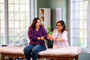 Hintli kadın fizyoterapist, modern klinikte yapılan detaylı istişare sırasında genç kadın hastaların bilek ağrılarını inceliyor, ortak değerlendirme ve rehabilitasyon alanındaki uzmanlığı vurguluyor.