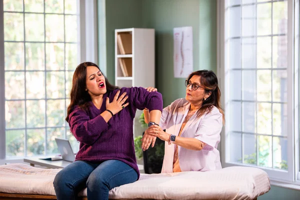 Hintli kadın fizyoterapist modern klinikte detaylı değerlendirme sırasında genç bayan hastaların omuz ağrılarını inceliyor, rotator manşet bakımı ve rehabilitasyon terapisinde uzman olduğunu gösteriyor.