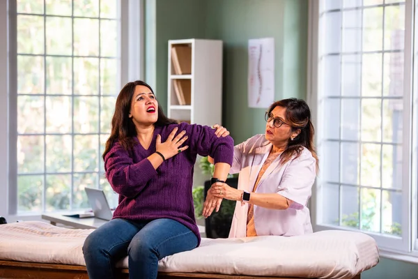 Hintli kadın fizyoterapist modern klinikte detaylı değerlendirme sırasında genç bayan hastaların omuz ağrılarını inceliyor, rotator manşet bakımı ve rehabilitasyon terapisinde uzman olduğunu gösteriyor.