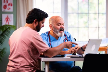 Hintli erkek doktor, deneyimli bir doktor olarak genç bir hastaya danışmanlık yaparken dizüstü bilgisayar kullanarak modern Hint kliniğinde OPD sağlık danışmanlığı hakkında konuşuyor.
