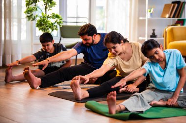 Hintli aile evde yog asana yapıyor Janu shirasana pratik yapıyor sakin bir sabah saatlerinde evde birlikte diz boyu yoga yapıyorlar modern savurgan bir yaşam alanında sağlıklı bir yaşam tarzını destekliyorlar.