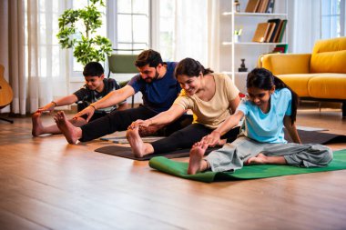Hintli aile evde yog asana yapıyor Janu shirasana pratik yapıyor sakin bir sabah saatlerinde evde birlikte diz boyu yoga yapıyorlar modern savurgan bir yaşam alanında sağlıklı bir yaşam tarzını destekliyorlar.