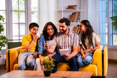 Hintli genç bir aile, evde eski fotoğrafları seyrederken birlikte mutlu anıları hatırlarken modern bir oturma odasındaki kanepede oturup gülüşmeler, duygusal aile anlarının tadını çıkarıyordu.