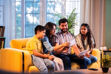 Hintli genç bir aile, evde eski fotoğrafları seyrederken birlikte mutlu anıları hatırlarken modern bir oturma odasındaki kanepede oturup gülüşmeler, duygusal aile anlarının tadını çıkarıyordu.