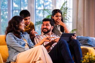 Evde tablet bilgisayar kullanan Hintli genç bir aile içeriği incelerken kanepede yan yana oturuyorlar ve modern, savurgan bir oturma odasında gülücüklerini paylaşıyorlar.