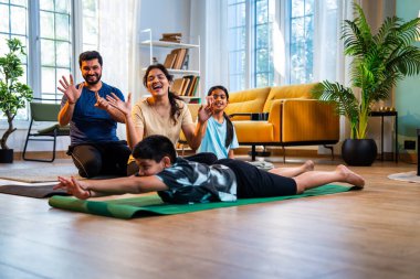 Tekne pozu veren Hintli bir çocuk yoga ya da evde denge çalışması yaparken aile sakin bir sabah saatinde modern, savurgan bir yaşam alanında sağlık hizmetlerini desteklerken evde denge egzersizi yapıyor.