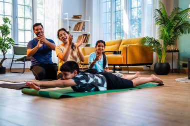 Tekne pozu veren Hintli bir çocuk yoga ya da evde denge çalışması yaparken aile sakin bir sabah saatinde modern, savurgan bir yaşam alanında sağlık hizmetlerini desteklerken evde denge egzersizi yapıyor.