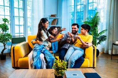 Evde çocukları gıdıklayan Hintli genç ebeveynler, neşeli oturma odasında kanepede otururken, mutlu baba ve anne içeride kızlarıyla gülüyorlar, neşeli aile hayatı.