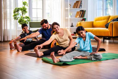 Hintli aile evde yog asana yapıyor Janu shirasana pratik yapıyor sakin bir sabah saatlerinde evde birlikte diz boyu yoga yapıyorlar modern savurgan bir yaşam alanında sağlıklı bir yaşam tarzını destekliyorlar.