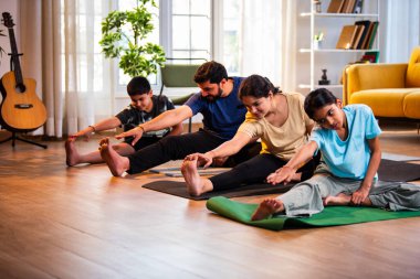 Hintli aile evde yog asana yapıyor Janu shirasana pratik yapıyor sakin bir sabah saatlerinde evde birlikte diz boyu yoga yapıyorlar modern savurgan bir yaşam alanında sağlıklı bir yaşam tarzını destekliyorlar.