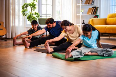 Hintli aile evde yog asana yapıyor Janu shirasana pratik yapıyor sakin bir sabah saatlerinde evde birlikte diz boyu yoga yapıyorlar modern savurgan bir yaşam alanında sağlıklı bir yaşam tarzını destekliyorlar.