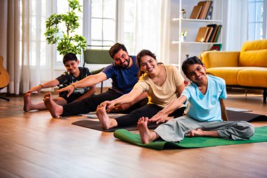 Hintli aile evde yog asana yapıyor Janu shirasana pratik yapıyor sakin bir sabah saatlerinde evde birlikte diz boyu yoga yapıyorlar modern savurgan bir yaşam alanında sağlıklı bir yaşam tarzını destekliyorlar.