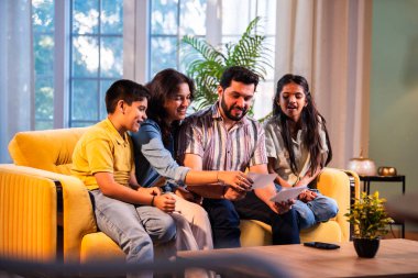 Hintli genç bir aile, evde eski fotoğrafları seyrederken birlikte mutlu anıları hatırlarken modern bir oturma odasındaki kanepede oturup gülüşmeler, duygusal aile anlarının tadını çıkarıyordu.