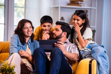 Evde tablet bilgisayar kullanan Hintli genç bir aile içeriği incelerken kanepede yan yana oturuyorlar ve modern, savurgan bir oturma odasında gülücüklerini paylaşıyorlar.