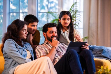 Evde tablet bilgisayar kullanan Hintli genç bir aile içeriği incelerken kanepede yan yana oturuyorlar ve modern, savurgan bir oturma odasında gülücüklerini paylaşıyorlar.