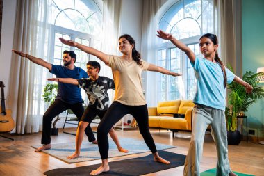 Hintli aile evde virabhadrasana yogası yapıyor. Savaşçı pozu veriyorlar. Sağlıklı vücut için evde poz veriyorlar.