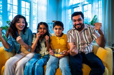Evde televizyon seyreden Hintli genç bir aile, evdeki rahat sinema gecesinde evde, modern bir oturma odasındaki koltukta oturup patlamış mısırın tadını çıkarıyorlar.