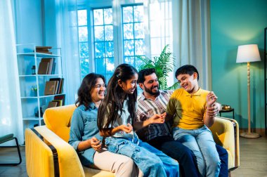 Evde çocukları gıdıklayan Hintli genç ebeveynler, neşeli oturma odasında kanepede otururken, mutlu baba ve anne içeride kızlarıyla gülüyorlar, neşeli aile hayatı.