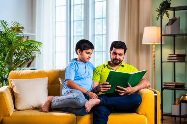 Hintli baba ve oğlu modern evdeki kanepede kitap okurken, baba okul bölümünü anlatıyor ve küçük çocuğa sıcak oturma odasında oturup aile eğitimi anlarını anlatıyor.