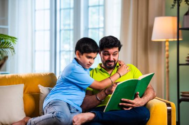 Hintli baba ve oğlu modern evdeki kanepede kitap okurken, baba okul bölümünü anlatıyor ve küçük çocuğa sıcak oturma odasında oturup aile eğitimi anlarını anlatıyor.