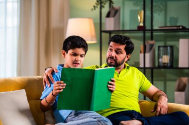 Hintli Asyalı oğlu babasıyla birlikte modern evdeki kanepede kitap okurken okul çocuğu özenle ders çalışıyor ve babası da rahat oturma odasında aile eğitimi anlarında öğrenmeyi destekliyor.