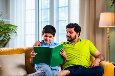 Hintli Asyalı oğlu babasıyla birlikte modern evdeki kanepede kitap okurken okul çocuğu özenle ders çalışıyor ve babası da rahat oturma odasında aile eğitimi anlarında öğrenmeyi destekliyor.