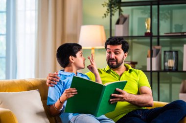 Hintli Asyalı oğlu babasıyla birlikte modern evdeki kanepede kitap okurken okul çocuğu özenle ders çalışıyor ve babası da rahat oturma odasında aile eğitimi anlarında öğrenmeyi destekliyor.